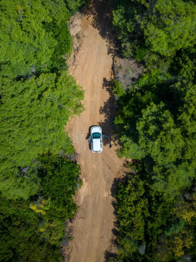 Aerial shot of a white car traveling on a dirt road surrounded by lush green trees in Skopelos, taken by a drone heading towards The Pig Lighthouse.